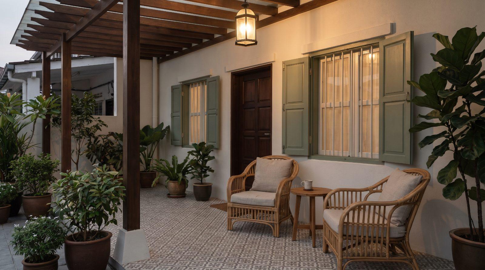The front porch of a Malaysian terrace house at dusk with two rattan chairs and a small table, warm porch light