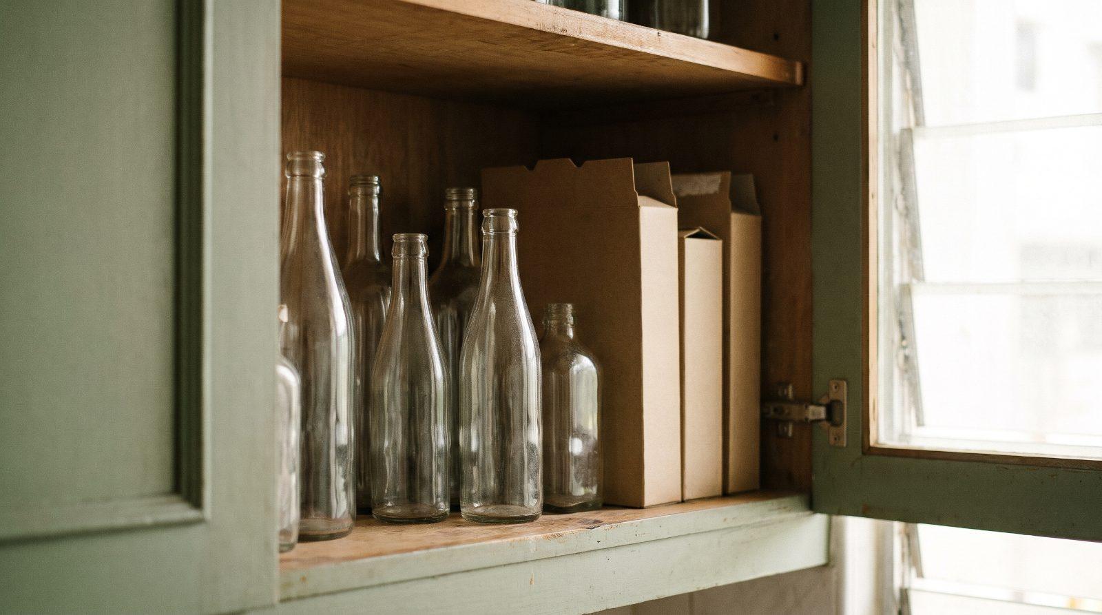 A kitchen cupboard with empty glass bottles half hidden behind cereal boxes, soft daylight